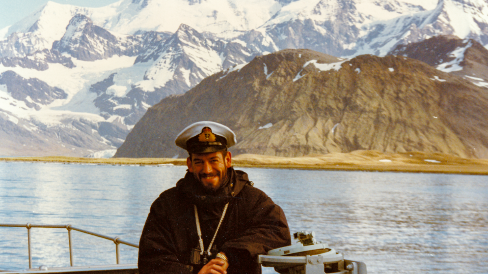 Commander Woods standing on a ship smiling with mountains behind him wearing a navy cap