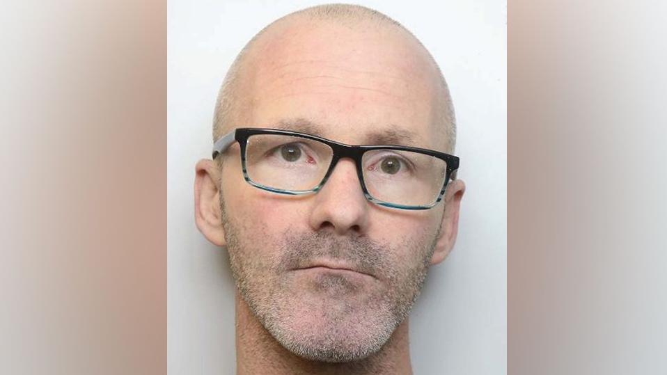 Gavin Collins who has a brown eyes, a bald head and grey stubble on his neck, chin and top lip and is wearing glasses with black frames  