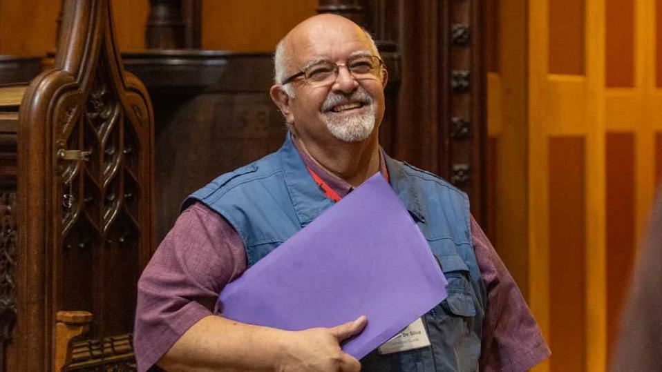 Stephen de Silva wears a maroon shirt and a blue gilet. He is carrying a purple folder. He has grey hair with a short grey moustache and beard and is balding. He is wearing clear-rimed glassed and is smiling.