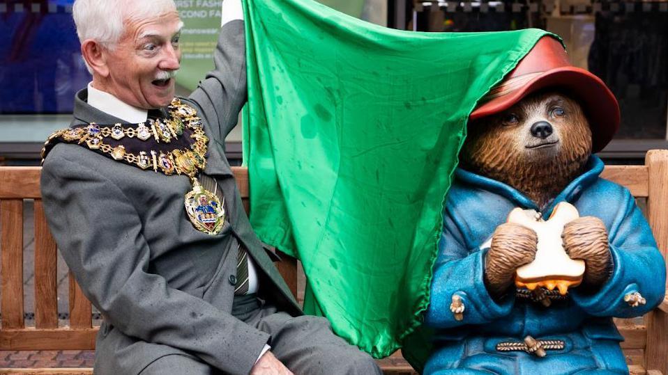 Mayor of Newbury Andy Moore - wearing a grey suit and with his ceremonial gold chain round his neck - sitting on the bench next to the statue of Paddington Bear and unveiling it from beneath a green, silk scarf