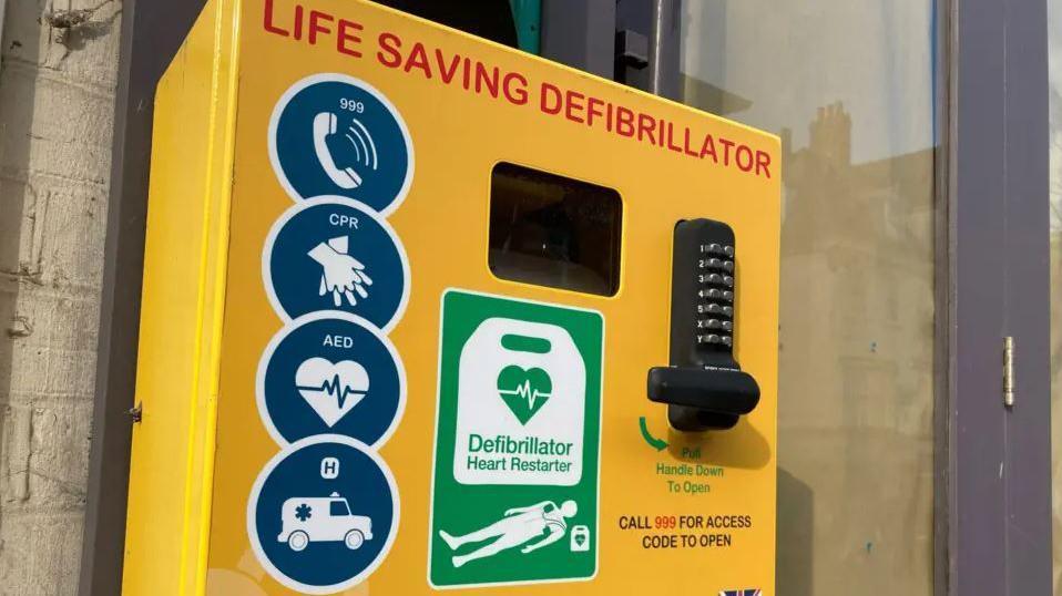 A defibrillator in a yellow box attached to a wall