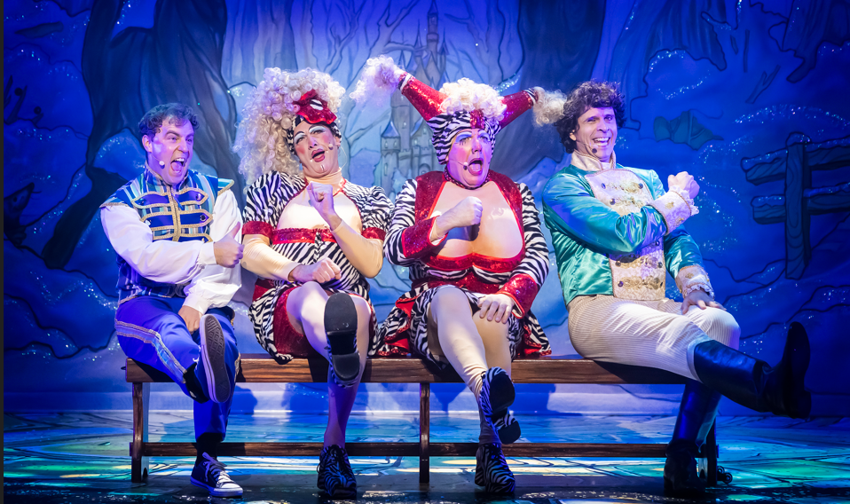 Four men sit on a bench on a stage. Two are dressed in blue waist coats, the two in the middle are dressed as pantomime dames with red dresses, stockings and white wigs.