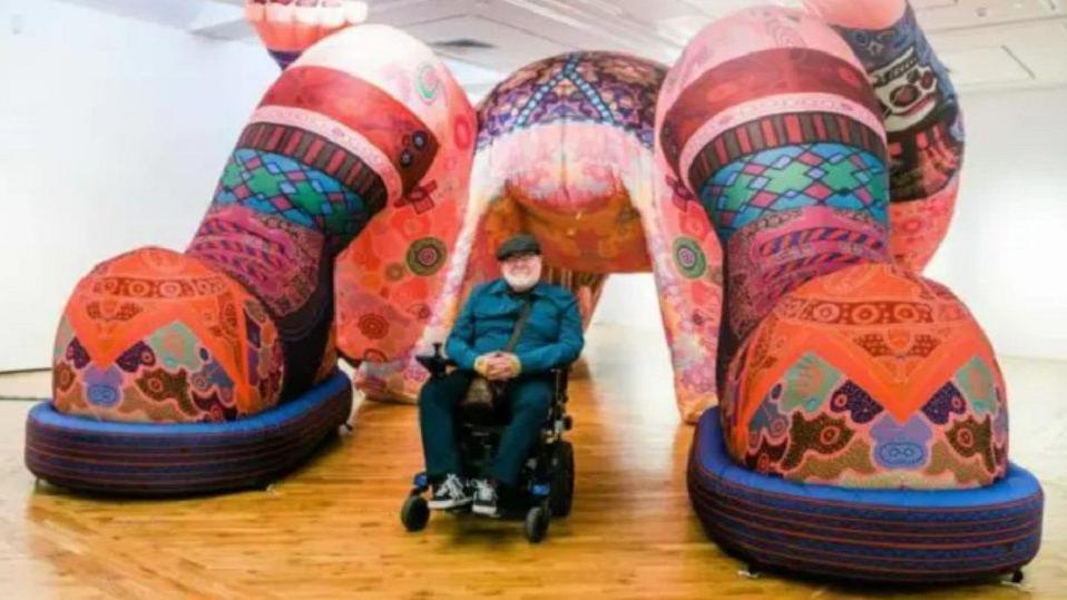 Artist Jason Wilsher-Mills sat in a wheelchair wearing a blue top, flat cap and blue trousers and trainers, with one of his large interactive sculptures filled with different colours.