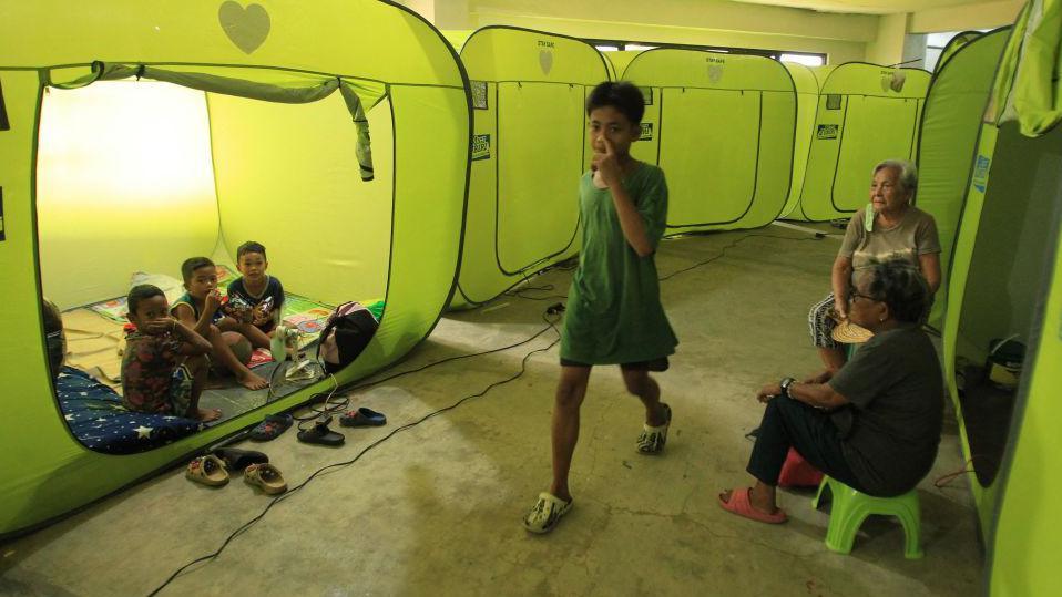 Two rows of green temporary shelters can be seen. A group of three children are sat in them whilst two older women watch over them - an older boy walks along a narrow concrete path between the two groups.