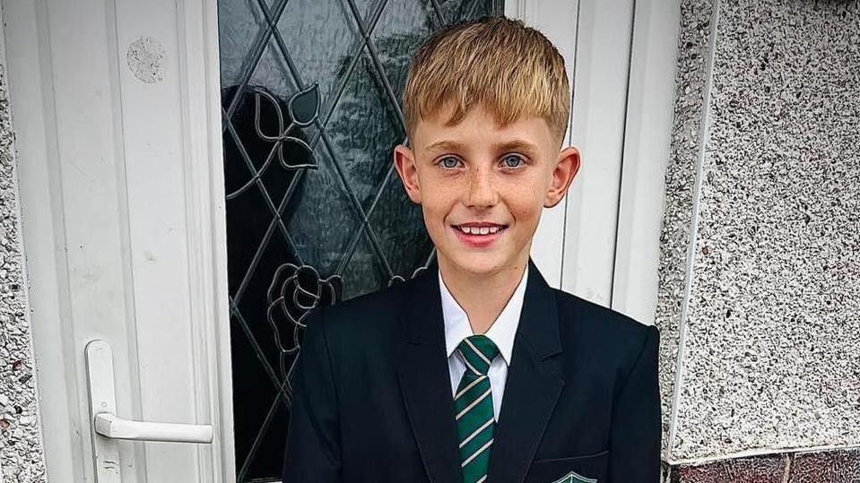 A boy with blond hair wearing a school uniform which comprises of a green and white striped tie and a blazer.
