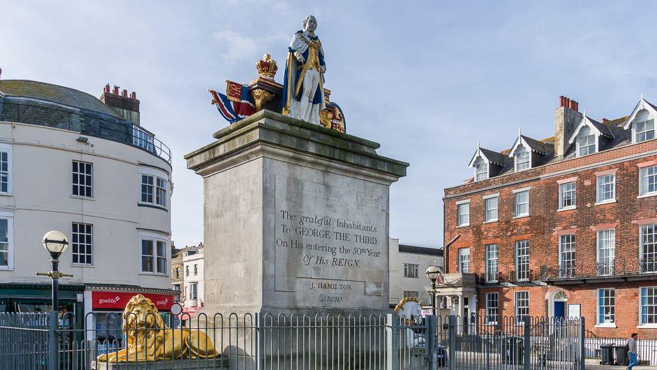 Looking up at the King George III statue on top of a large stone plinth. The painted statue depicts the king standing between a crown and a pile of books. At the base of the plinth is a gold lion and a white and gold unicorn.