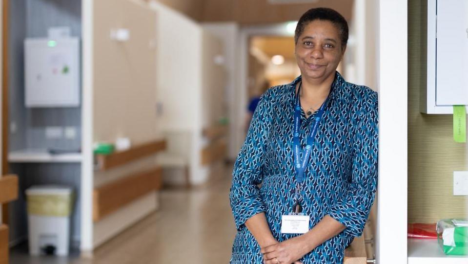 Susan Hamilton wearing a blue and black patterned dress and a blue lanyard. She is leaning against a wall in a hospice building, with medical supplies dotted around. She has short black hair and is smiling at the camera with her hands folded.