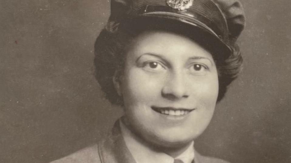 A black-and-white photograph of a young woman in uniform. She has dark brown hair and is smiling at the camera.
