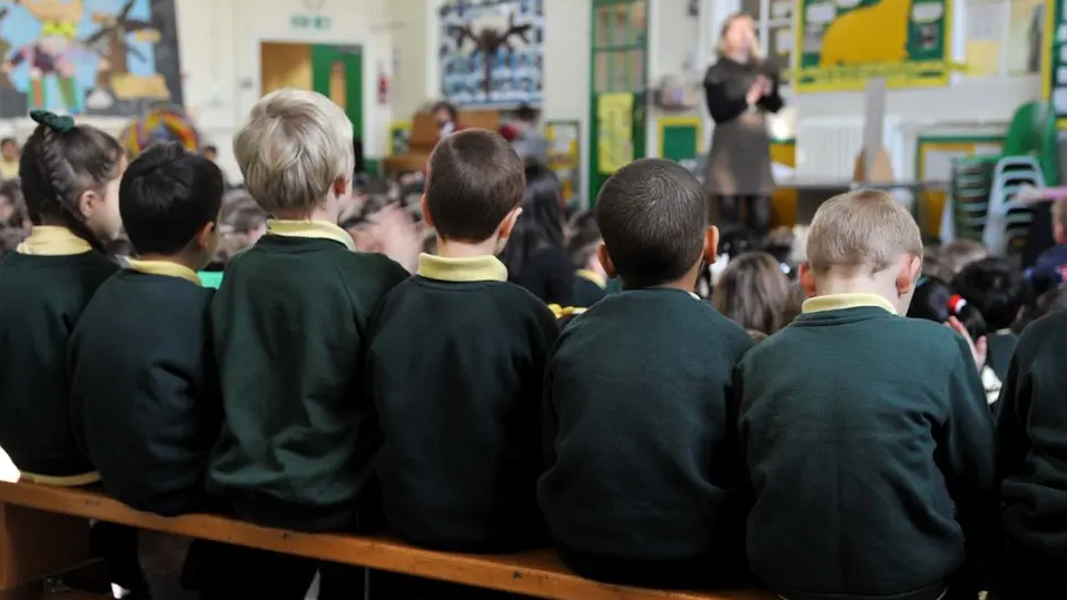 Six children sit in a row in class with their backs to the camera. They are wearing a green uniform and yellow shirts. 