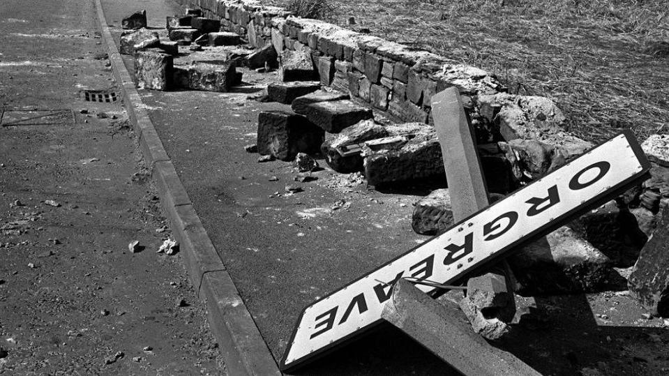 A black and white image of a twisted sign, felled concrete posts and a broken wall following violence outside the coking plant in Orgreave, South Yorkshire.