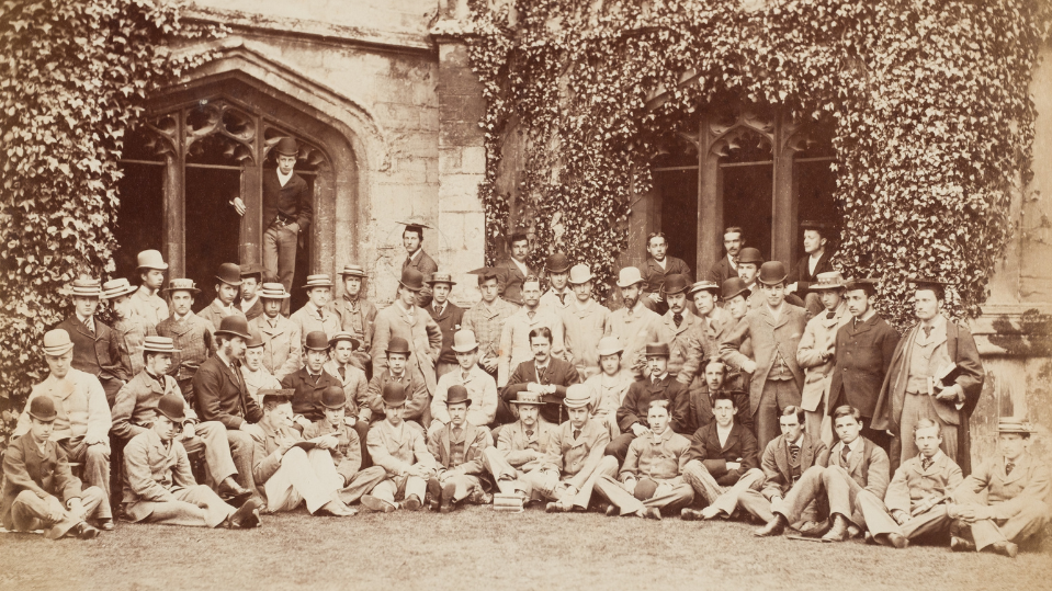 A black and white group photo taken in the cloisters of Magdalen College, Oxford, in 1876. The men in the image are all well dressed.