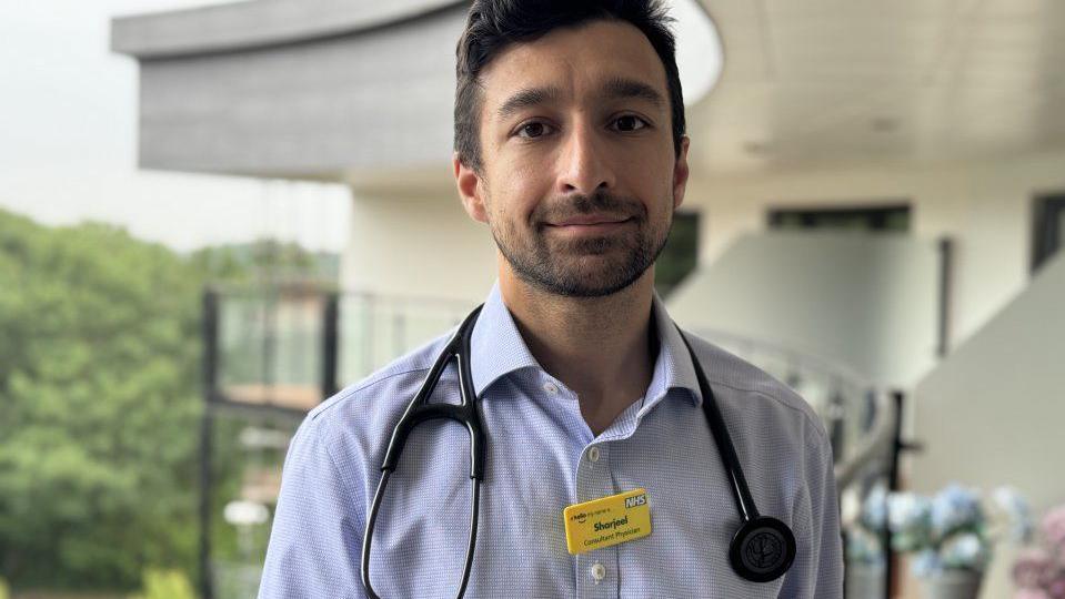 A doctor with dark hair and a beard is wearing a yellow name badge. He is standing outside with a building in the background.
