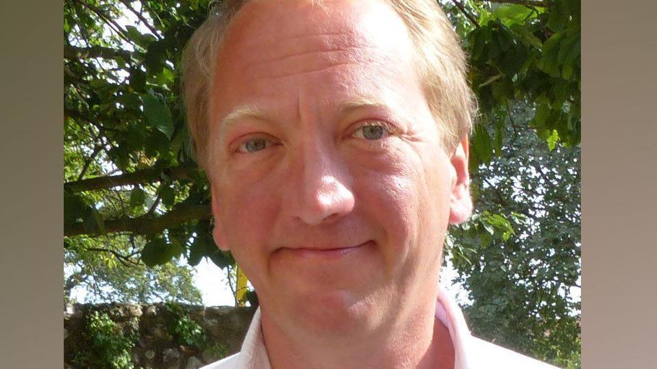 Adrian Marsden with smiling at the camera, with short fair hair, and trees in leave behind him