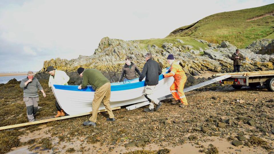 People push the white and blue boat down a ladder and wooden plank on a rocky beach.