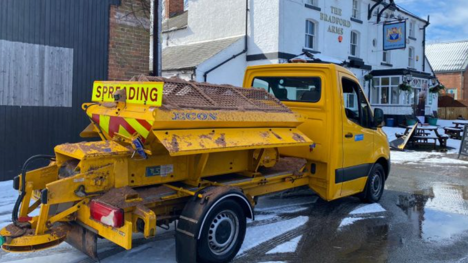 A yellow van with a spreader on the back, outside a pub in the snow