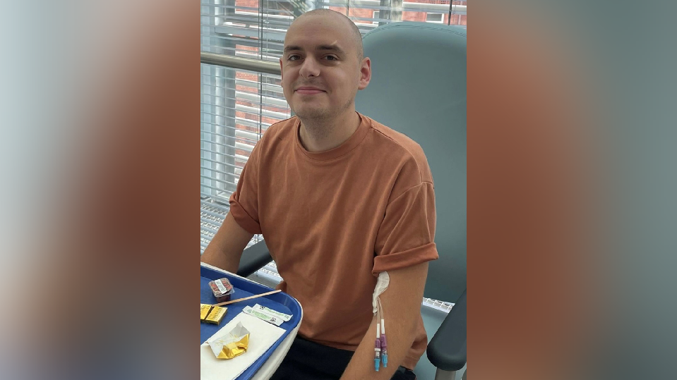A man with a shaved head and peach coloured T-shirt sits with a catheter in his left arm.