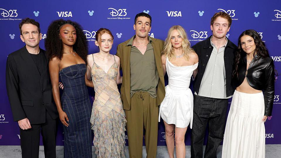The cast of the new X-Men movie all standing up posing for a picture. Jake is in all black, Maya is in a dark blue dress, Sadie is wearing a dress with a rectangle pattern on it, Christopher is wearing an olive blazer and trousers, Samantha is wearing a white dress, Kit has a black jacket on with a stripy shirt on underneath, Inde is wearing a black leather jacket with a white skirt.