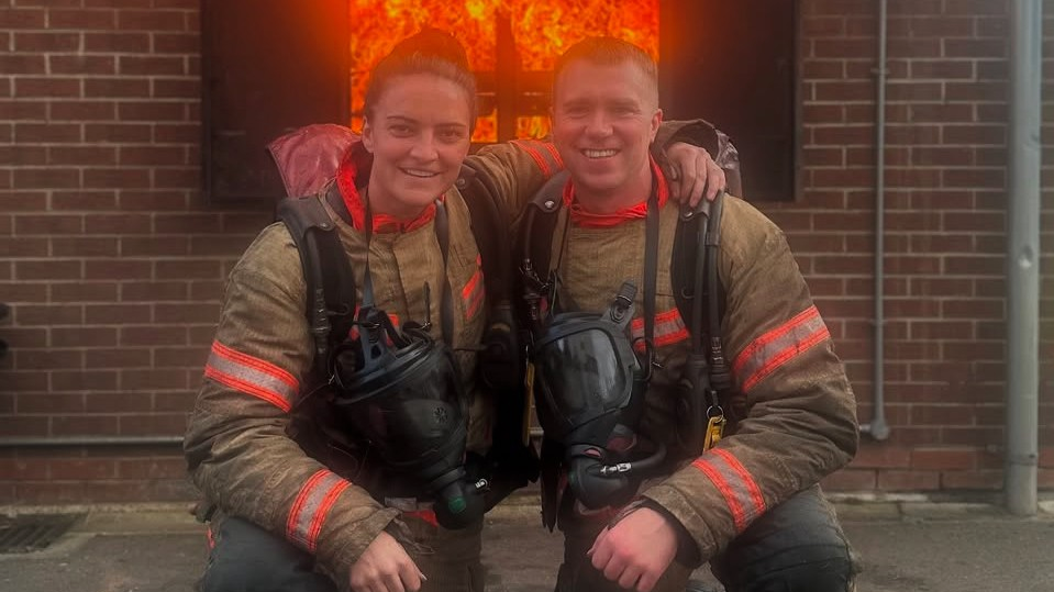 Jess Sigsworth and a male firefighter outside a building with flames in the background