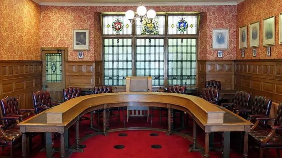 The Legislative Council Chamber, which has a red carpet and a brown desk in the shape of the horse shoe. Leather seats are positioned around it and the walls have wooden panelling and pictures hung on them.