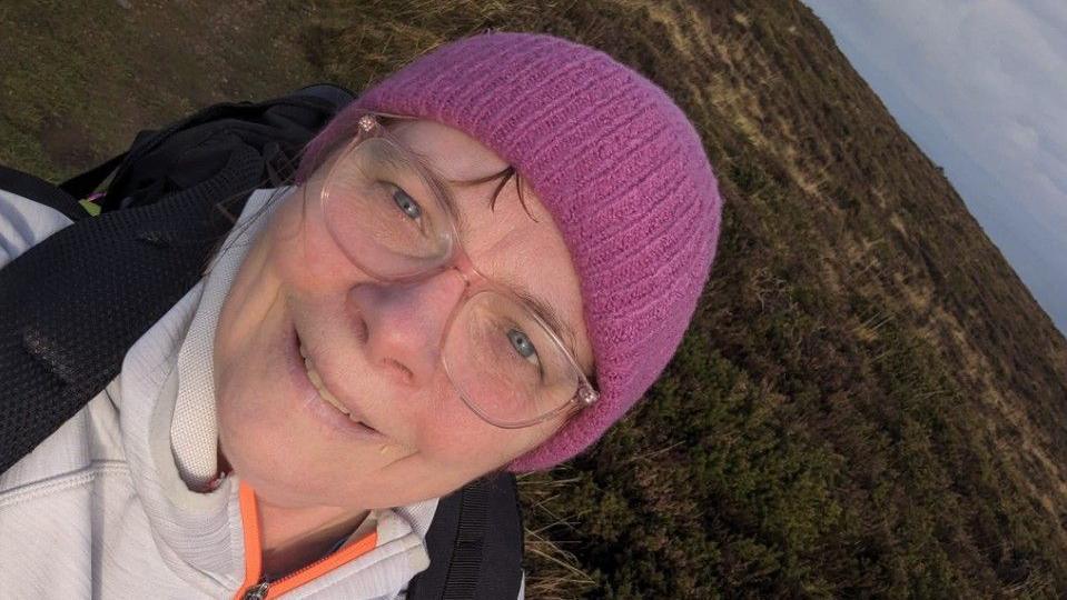 Liz has glasses and a pink hat and you can see the coastal vegetation behind
