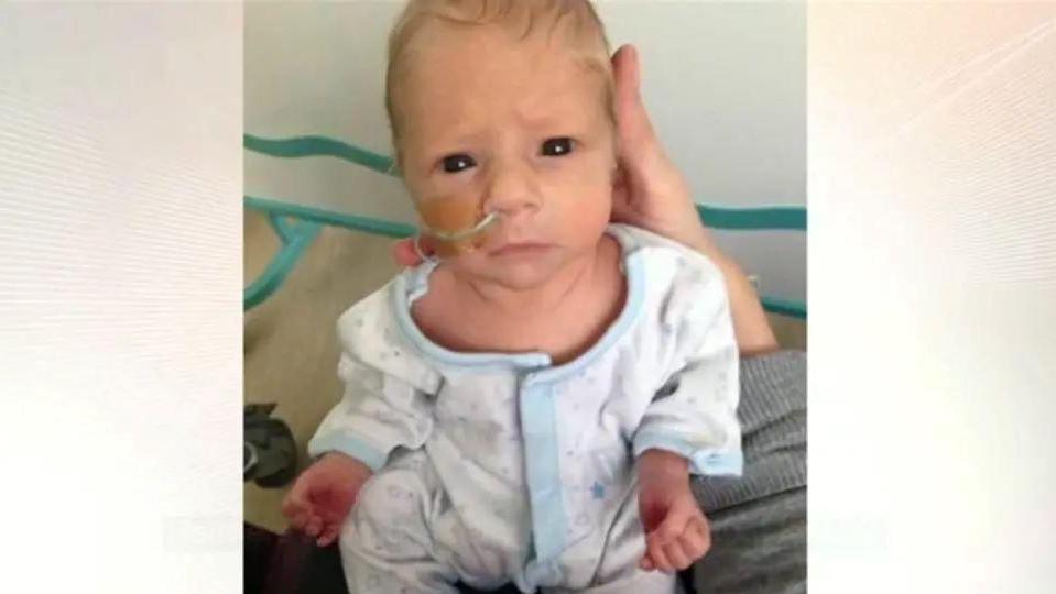 A new born baby is held up to the camera. He wears a white and blue baby grow and has a nasal tube. 