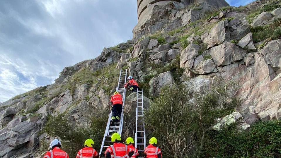 Five firefighters wearing white and yellow helmets stood at the bottom of two ladders. There is one firefighter climbing up one of the ladders, and another on the cliff face. There are grey rocks on the cliff face.