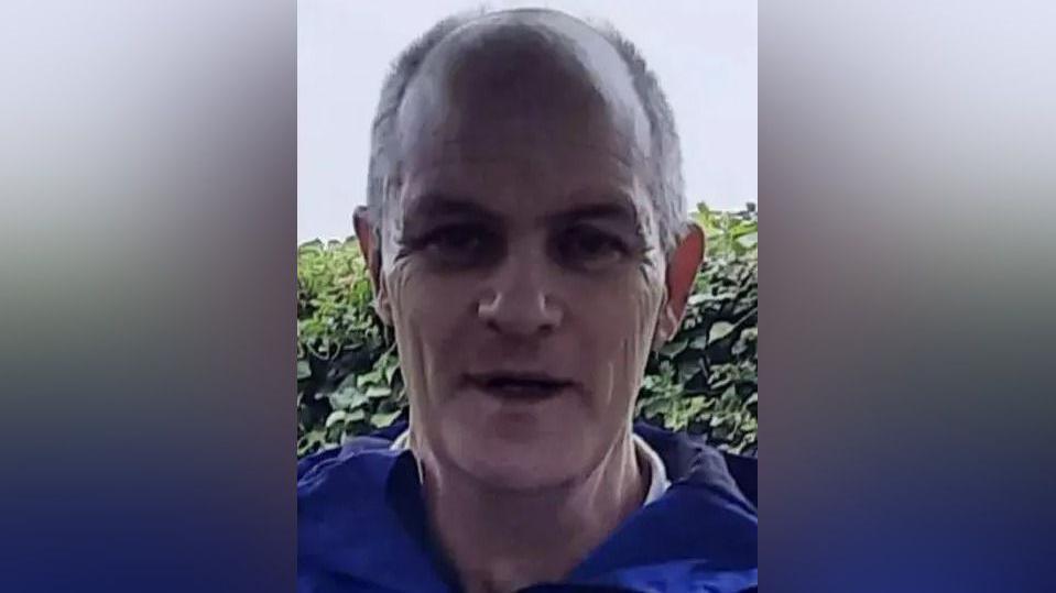 Thomas Tierney looks at the camera. He is bald with grey hair on the sides and is clean shaven. He is wearing a blue jacket with a green hedge behind him