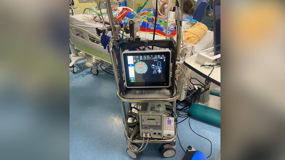 Extracorporeal Membrane Oxygenation machine, a tall machine with wires coming out of it. Behind it is Freddie in a hospital bed with a coloured blanket covering him