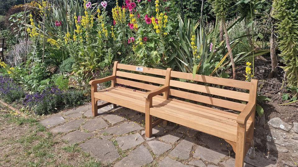 A shot of a new garden bench with lots of colourful flowers behind it 