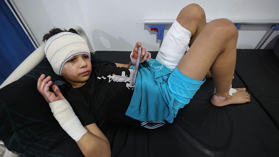 A young child, with a heavily bandaged head, right arm and left thigh, rests on a hospital bed after treatment in Gaza. He is wearing a black T-shirt and blue shorts and is holding a phone or small electronic tablet in his hand.