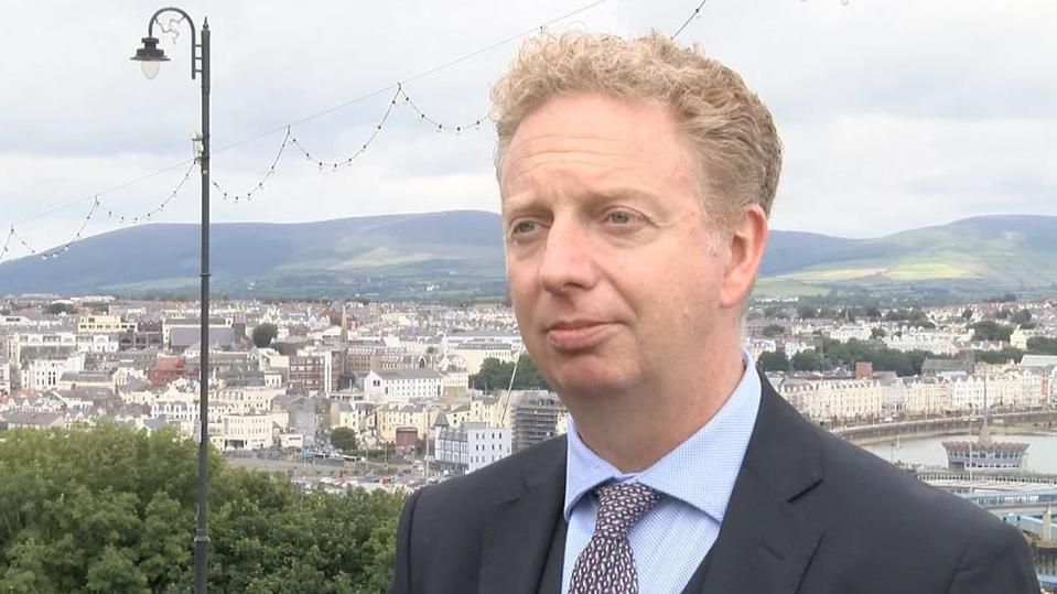 Alex Allinson, who is wearing a grey suit, blue shirt and purple patterned tie. The white buildings of the centre of Douglas and the sweep of the bay can be seen in the distance behind him, with rolling hills on the horizon.