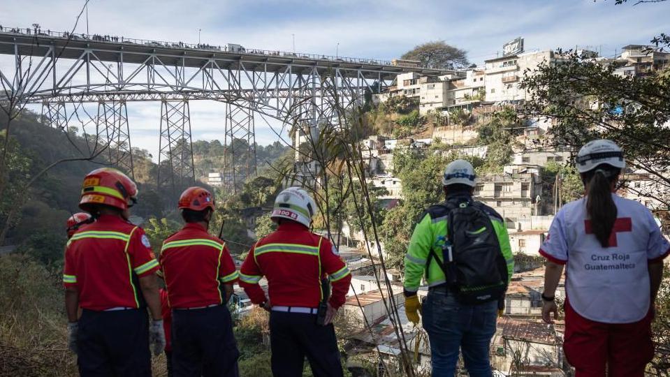 Rescue workers wearing uniforms and helmets observe the bottom of a ravine into which a bus fell on the outskirts of Guatemala City. The bridge can be seen in the background, towering above the deep ravine. 