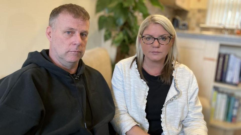 Jenny and Allyn Condon sit together on a sofa looking at the camera. They both have very serious expressions on their faces. He is wearing a black hooded top and she has a white cardigan and black top on