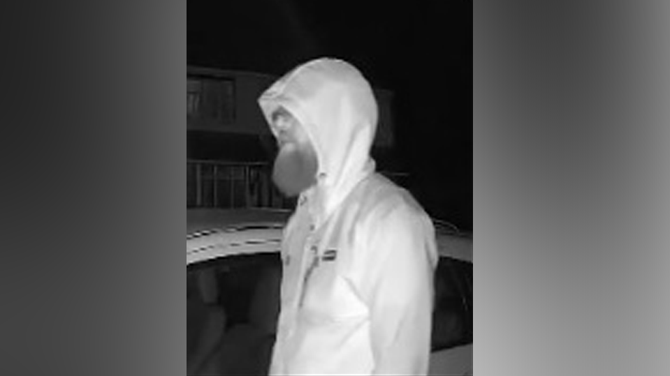 A CCTV black and white image of a man with a long beard, wearing a grey sweatshirt with the hood up. 