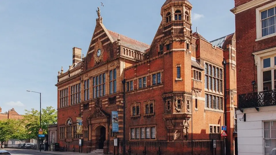 The old and ornate red-brick building stands on a main road in the city. It has a small clock above its entrance.