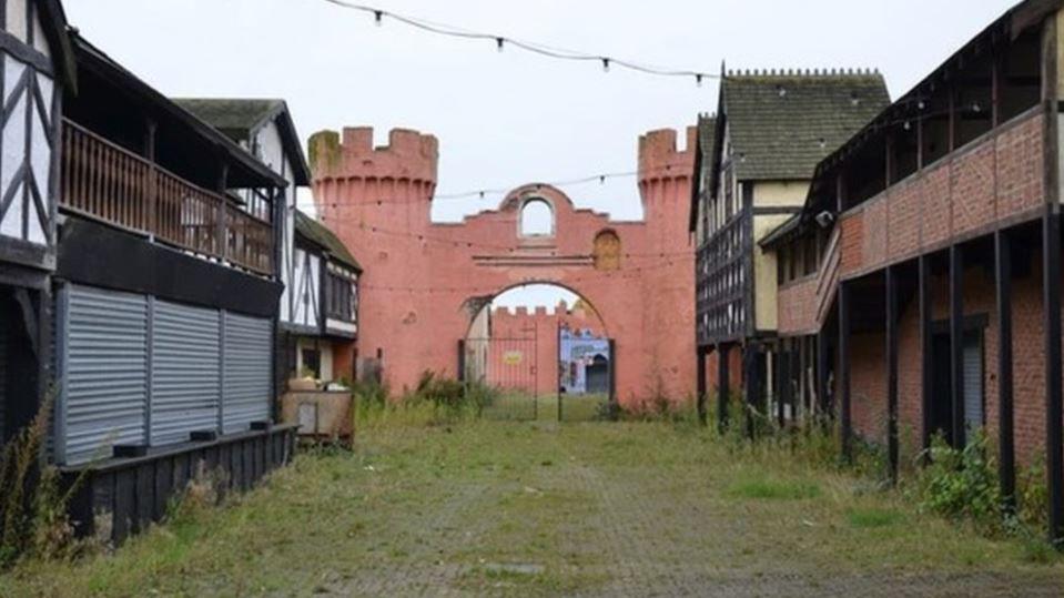 Overgrown area on site of the derelict Pleasure Island with two castle-style pink turrets and an archway. In the foreground are mock-Tudor buildings.