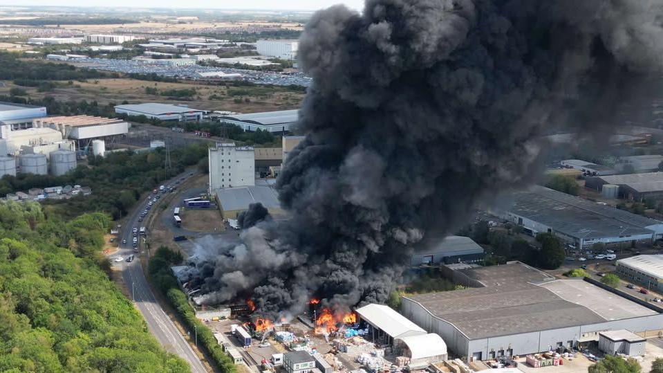 A large fire at an industrial estate, there are flames and a large thick plume of black smoke coming out of the building. There are cars parked nearby. 
It is an overheard view of the site.