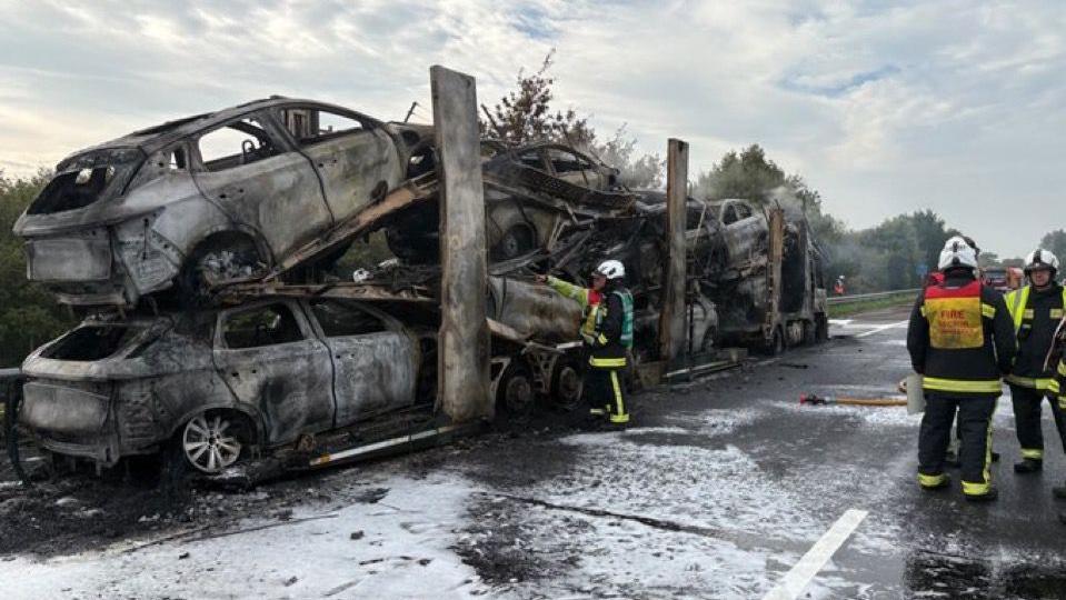 A number of cars affected by fire damage stacked on top of one another on the HGV. There are fire crews at the scene. 