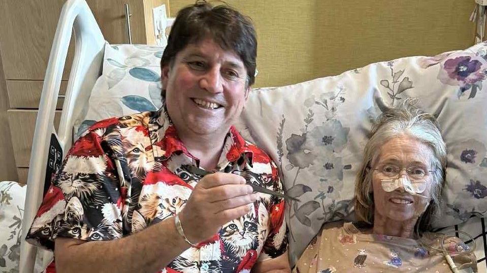 John and Rowena Owen sit in a large hospital type bed. Rowena has a tube going into her nose, held in place by micropore tape. She is wearing a top with cats on while John wears a shirt with cats on. The are both smiling. Rowena is looking into camera but John is looking slightly off camera.