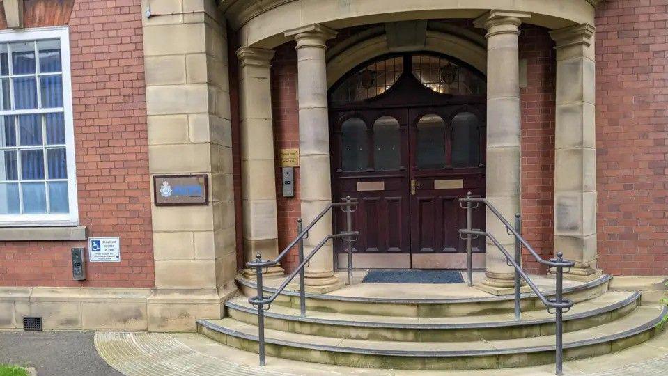 The entrance to a police building. Visible are a set of oak front doors, with steps leading up to them and stone pillars on either side.