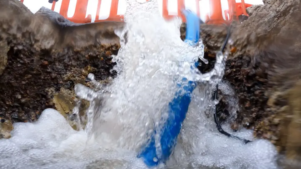 Water is gushing into a hole in a road. The image is close up from inside the hole and looking upward. There is a blue pipe half in and half out of the hole. Above on the road, you can see orange fencing around the hole.