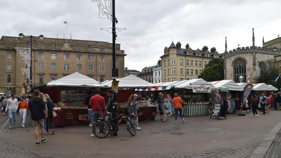 The photo is of the Cambridge market. The sky is grey and cloudy and there are buildings of various colours in the background. On the ground the camera is focused on the market stalls which have white, blue, yellow or green striped tarp covers atop. The stalls are on cobbled flooring and around the market is a smoother concrete path. There are customers walking around the market, one with a bike, browsing the stalls. Signs for a olive stall and a coffee stall can be seen clearly from the camera angle.