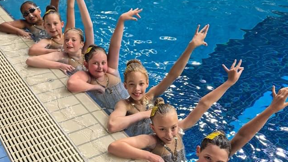 Young girls perched on the side of a swimming pool, in the water, with arms raised.