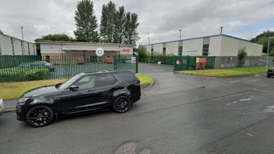 a black car sits outsied the green gates of a royal mail delivery office