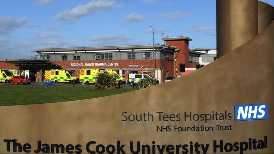 An entrance sign for The James Cook University Hospital is in the foreground of the photograph. In the background, a number of ambulances are parked outside the hospital's Regional Major Trauma Centre.