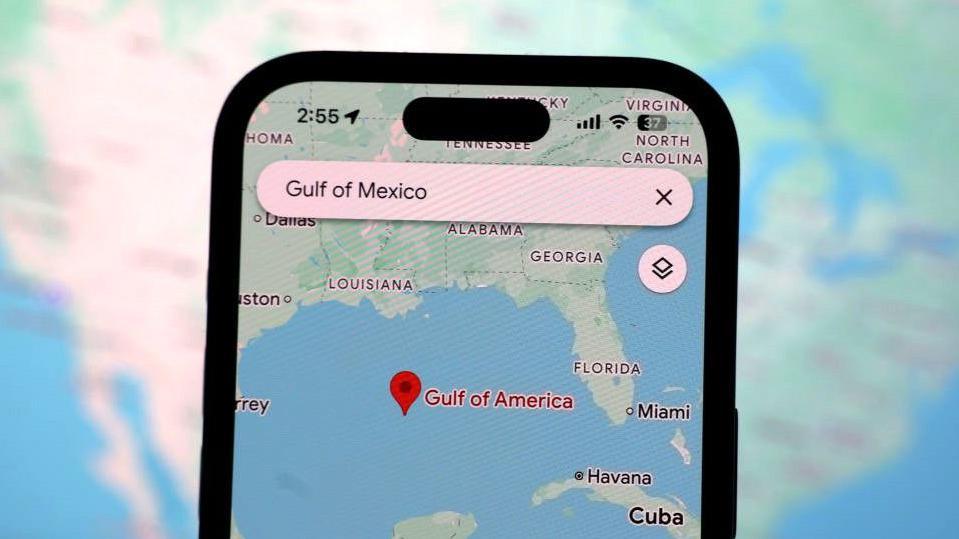 An iPhone screen displays a Google Maps app search for "Gulf of Mexico", with a pin on the Maps location showing the name "Gulf of America" below it. A map of North America is shown in a blurred backdrop behind it.