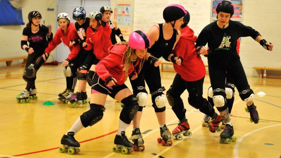 Roller derby players wearing helmets and protective gear train indoors on a sports court. Several skaters in red hoodies and black outfits form a line while others practise blocking and manoeuvres.
