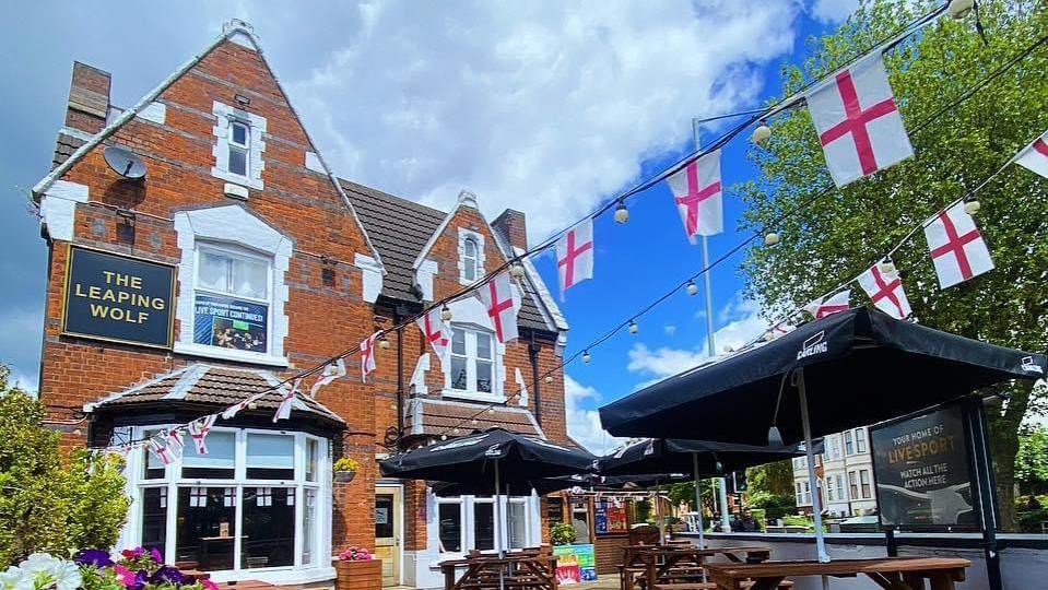 A red brick pub with a black sign with gold text which says the leaping wolf.
There are England flags on bunting outside the building