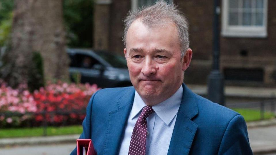 Simon Hart walking up Downing Street with a tight-lipped expression and carrying a red file under his arm.