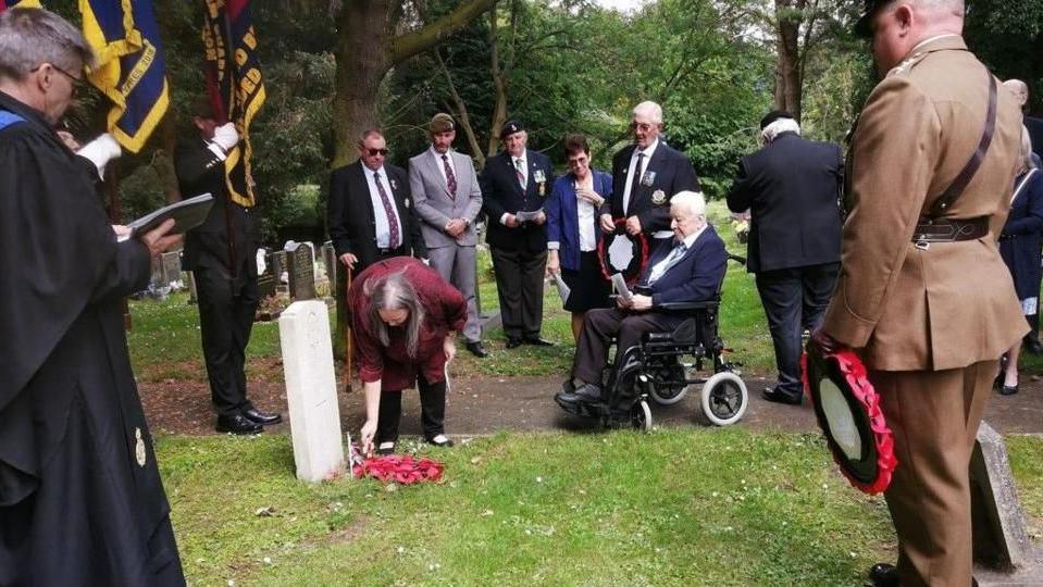 A group of people stand around a Commonwealth war grave. A woman is laying a wreath, and a soldier stands by holding one. 